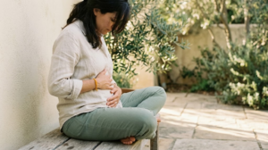 A person seated from the side on a low outdoor bench, head lowered, taking a slow deep breath with both hands resting on their belly.