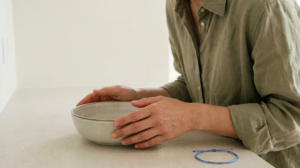 A person in a sage linen shirt leaning over a shallow ceramic bowl of cold water on a pale table.