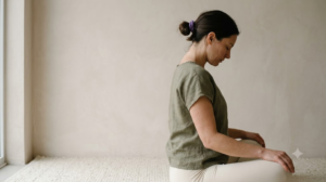 A person in an olive linen top seated on a mat by a plain wall, slowly rolling their shoulders back.