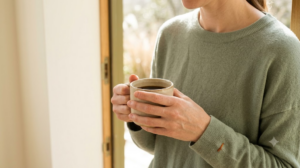 A person in a sage jumper holds a plain cup of coffee with both hands in a bright open doorway.