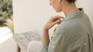 An over the shoulder view with the face turned away on a low bench outdoors, a person tapping their fingertips lightly on their collarbone in bright diffuse daylight.