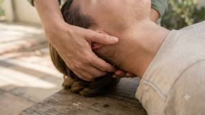 A person lies face-up while another person's hands gently cradle the back of their neck in a calm, softly lit room.