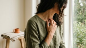 A person humming with two fingers resting lightly at the base of their throat beside a bright window.