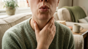 Close crop of a persons mouth, neck and shoulders as they exhale slowly with one hand resting at the side of their neck in a deep armchair.