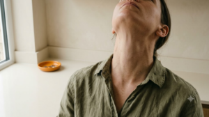 A photograph of a person with head tipped back gargling a mouthful of water, face out of frame, in a calm cream room.