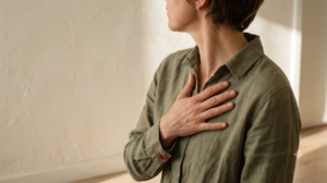 A person in soft sage clothing breathing slowly with one hand resting flat on their chest, face turned away, in warm morning light.