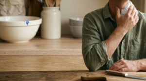 A person holding an ice pack against the side of their neck at a wooden kitchen counter in soft window light.