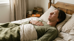 A person lying back on rumpled bed linen letting a long slow breath out with one hand on their upper chest.