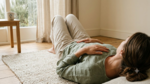 A person lying on their back on a rug with one hand on their chest and one on their belly in soft natural light.