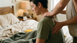 A practitioner&rsquo;s hands working on a seated person&rsquo;s shoulder and upper back in a warm bedroom