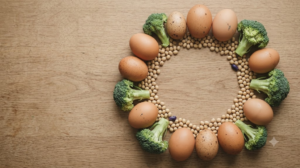 Whole eggs, soybeans and broccoli florets arranged in a loose ring on a plain oak board, shot from above.