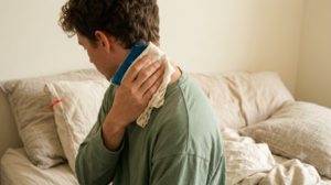 A photograph seen from behind of a person holding an ice pack against the side of their neck in soft daylight.