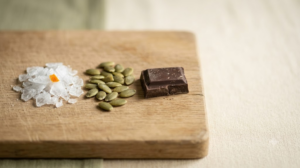A still life photograph of a small heap of magnesium flakes with pumpkin seeds and a piece of dark chocolate on a plain pale surface.