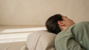 A person in a sage top resting on a linen cushion with fine acupuncture needles placed in their outer ear in warm afternoon light.
