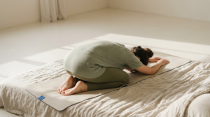 A photograph of a person resting in child’s pose on a yoga mat, face out of frame, in soft daylight.