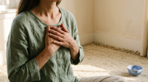 A close view of hands and a collarbone from just above, both hands resting over the heart while breathing slowly, on a woven rug in warm afternoon light.