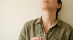 A person cropped at the chin tilts their head back to gargle water from a glass against a plain plastered wall.
