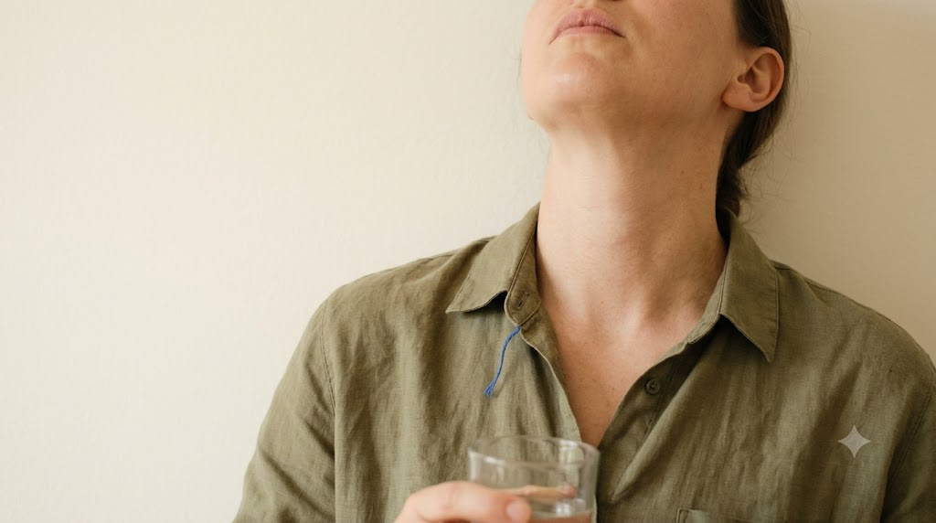 A person cropped at the chin tilts their head back to gargle water from a glass against a plain plastered wall.