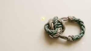 Overhead clay knot of sage and putty cords loosening into a ring, with a single golden spark suspended above it.