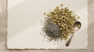 Overhead still life of a small heap of dried lavender buds and chamomile flowers with a worn spoon beside them on cream paper.