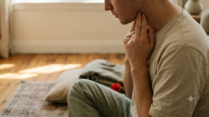 A seated person presses two fingertips into the side of their neck in low morning sunlight.