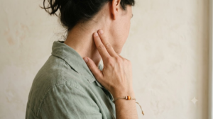 A photograph of a person tracing two fingers down the side of their neck, face out of frame, in soft daylight.